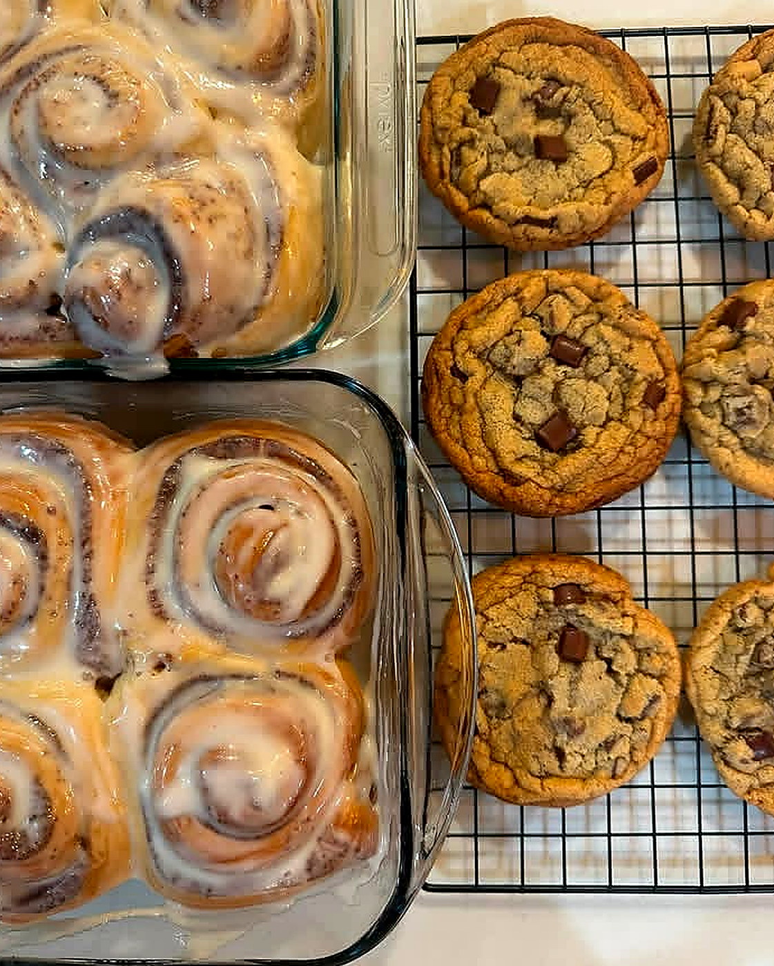 Fresh iced cinnamon rolls in glass pans beside chocolate chip cookies on a cooling rack